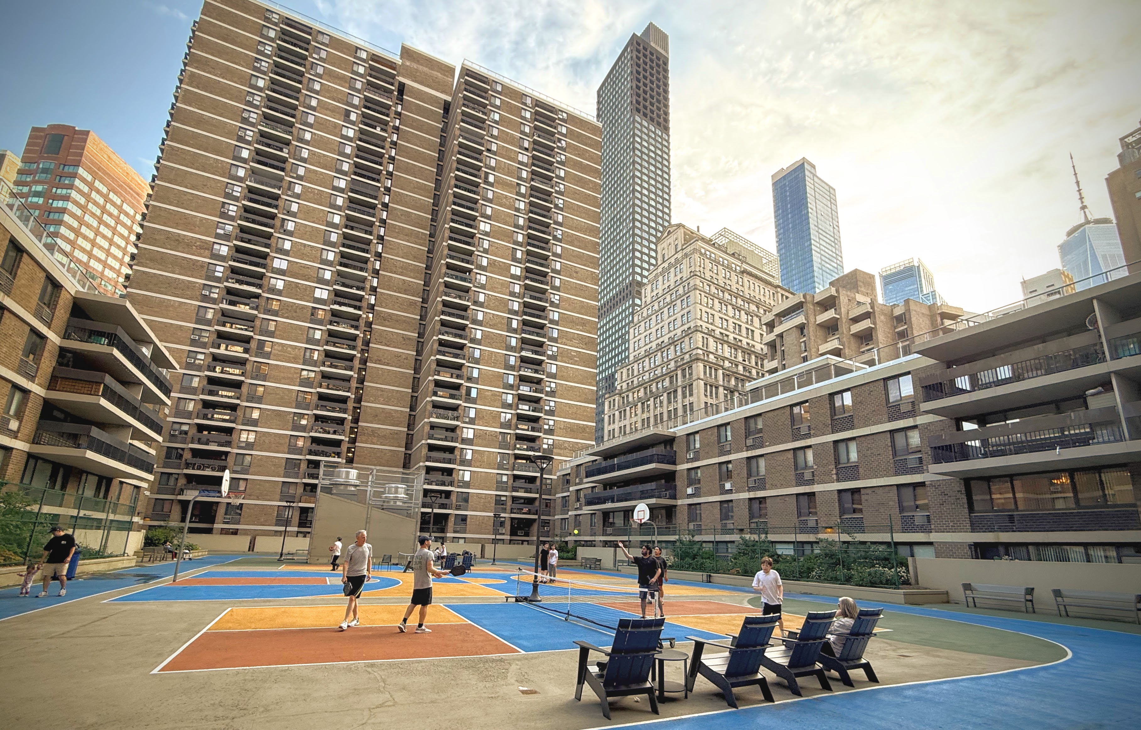 Residents playing on a Southbridge sports court at golden hour, with Adirondack chairs courtside and the Southbridge towers and Lower Manhattan skyline behind